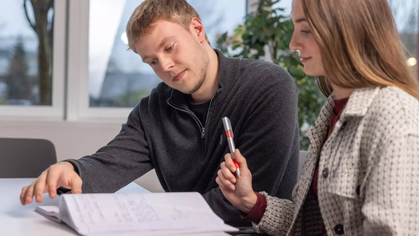 Ein Mann und eine Frau schauen in ein Notizbuch, die Frau hält einen Kugelschreiber, beide lächeln und sitzen in einem modernen Büro