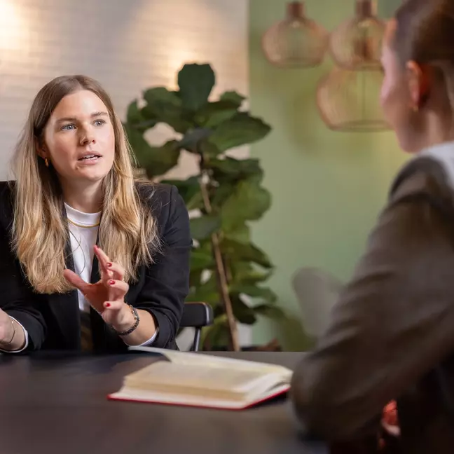 Zwei Frauen im Gespräch in einem modernen, freundlich gestalteten Büro. Es liegt eine aufgeschlagene Mappe auf dem Tisch 