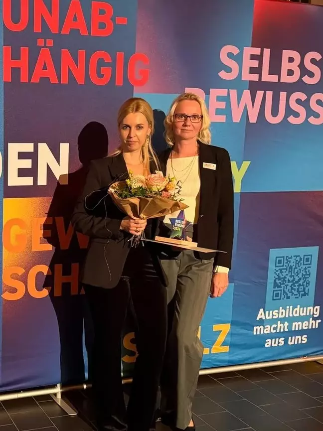 Zwei Frauen stehen vor bunter Rückwand, eine hält einen Blumenstrauß und eine Trophäe nach einer Ehrung in der Hand.