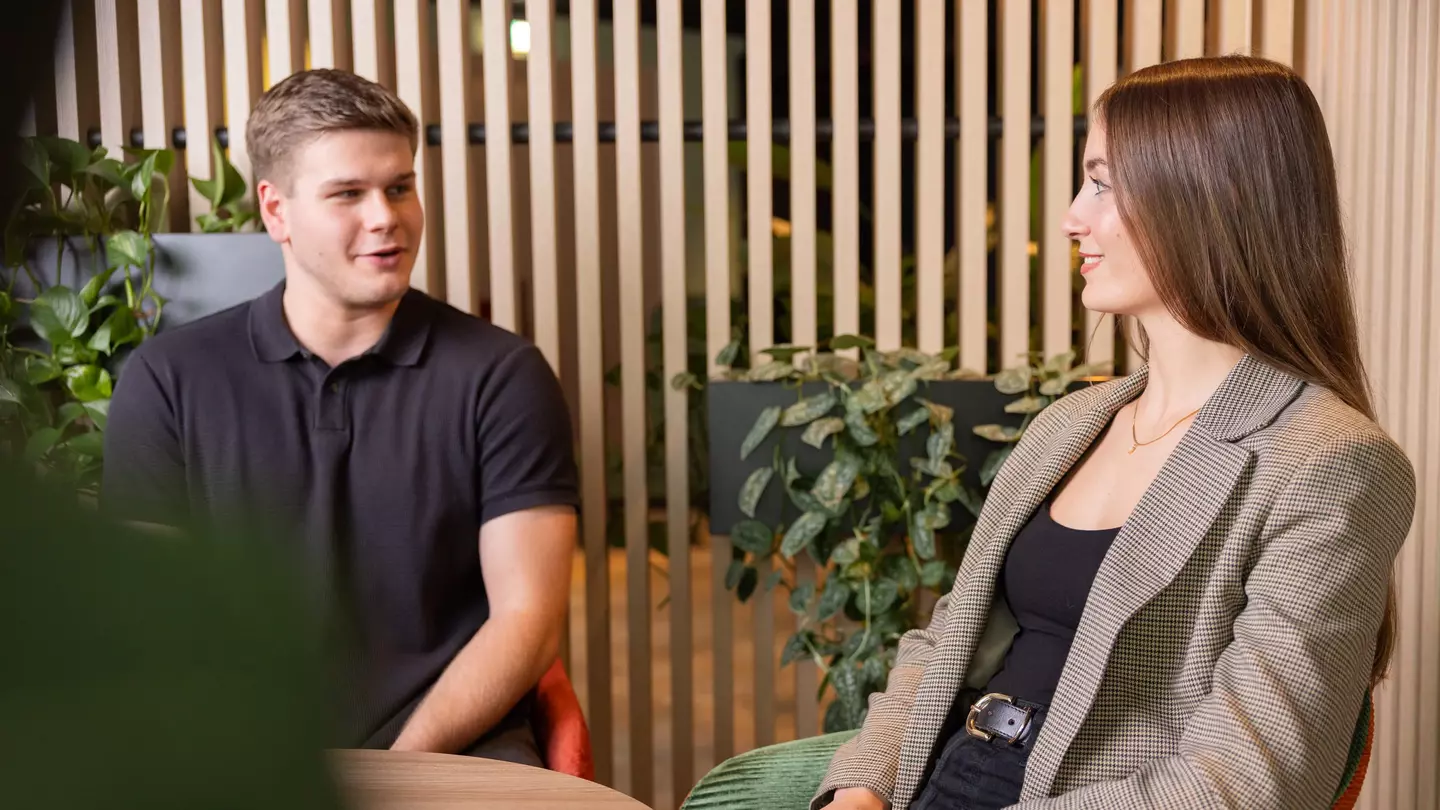 Zwei Mitarbeitende sitzen an einem Tisch und reden miteinander, sie sitzen in einem modernen Büro und lächeln