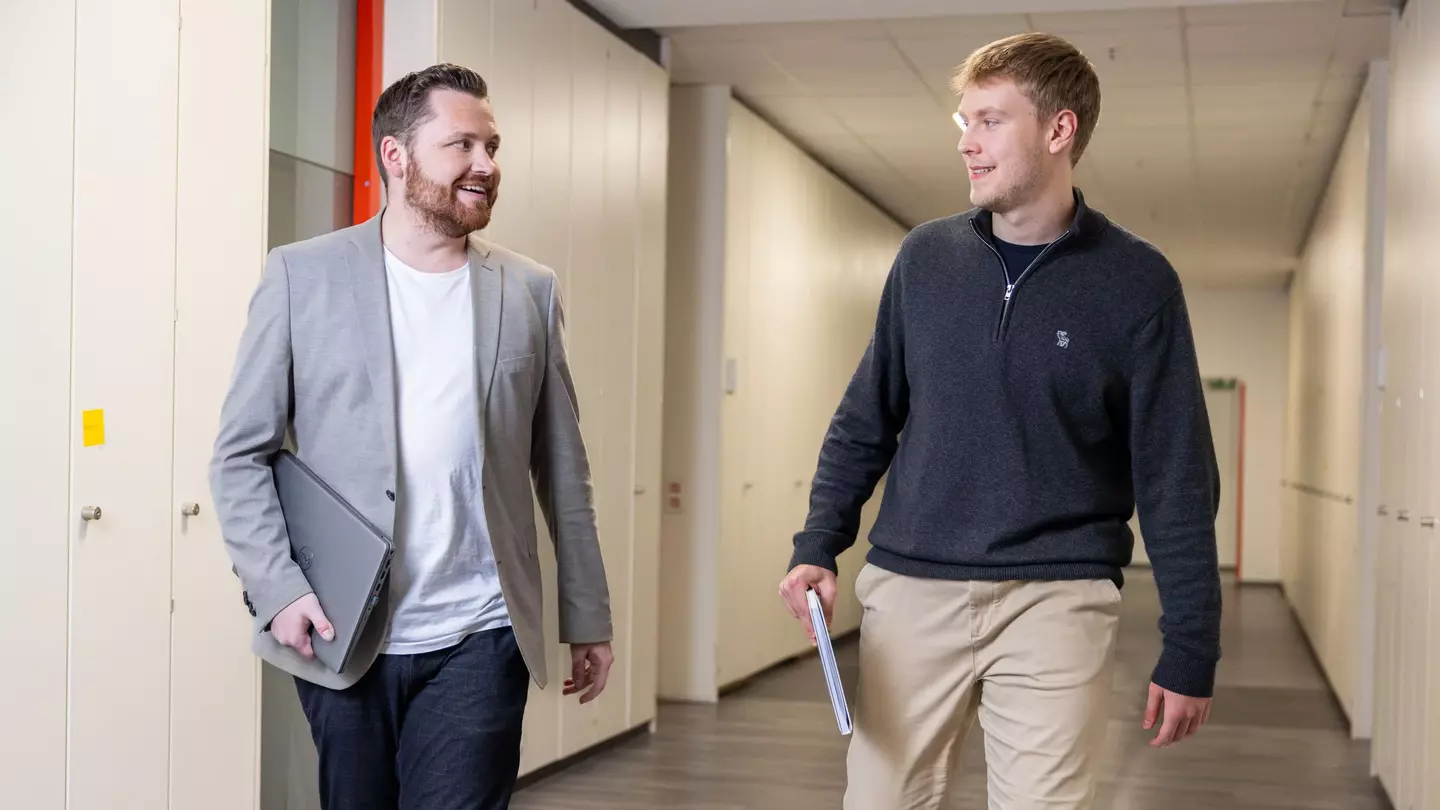 Zwei Mitarbeiter gehen lächelnd über einen Flur. Einer hält einen Laptop und der andere ein Notizbuch, sie schauen sich an