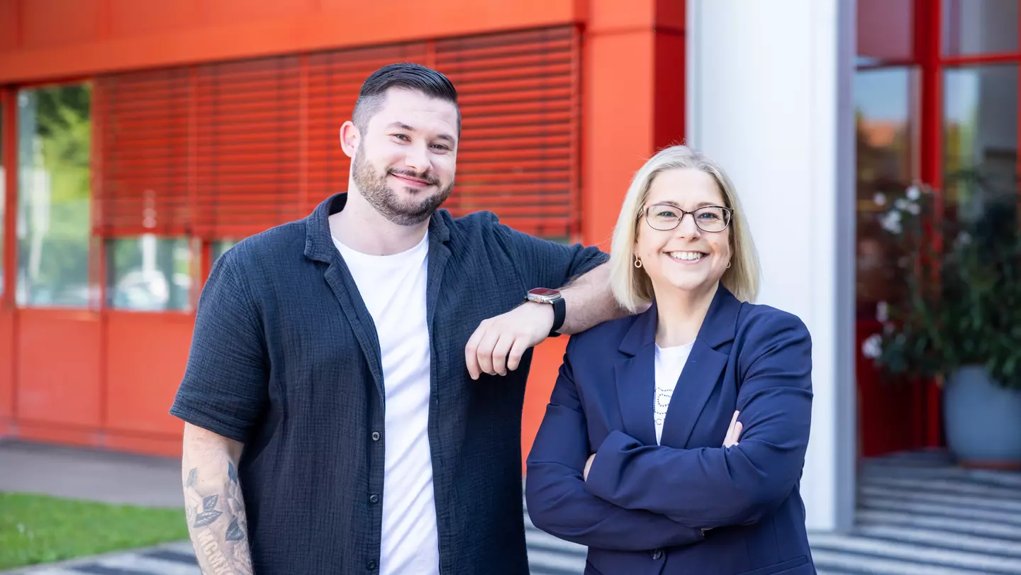 Zwei Personen stehen vor einem Gebäude im Außenbereich und blicken freundlich in die Kamera.