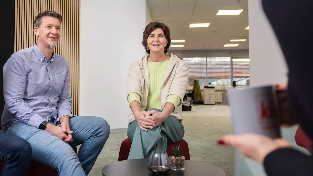 Ein Mann und eine Frau sitzen in einem modernen Bürobereich, lächeln und unterhalten sich mit einer Person, die eine „kik“-Tasse hält.