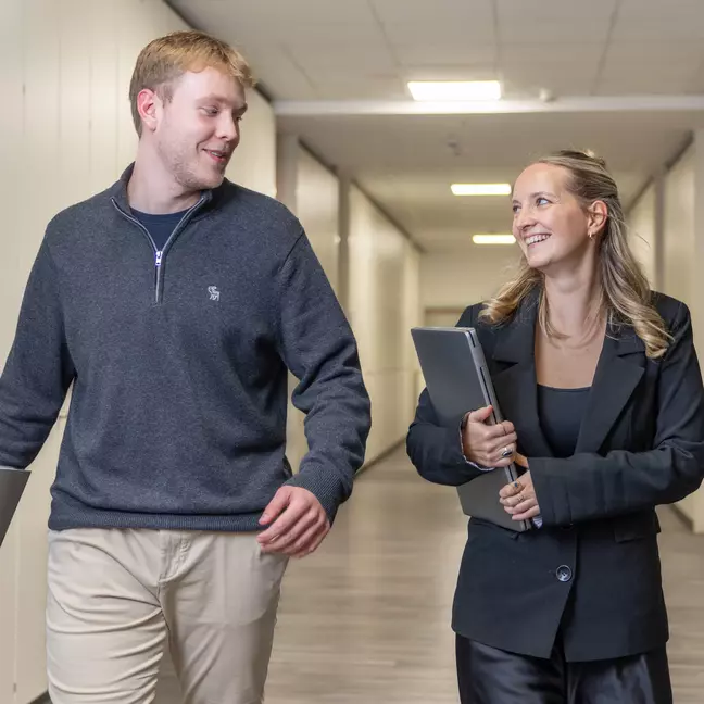 Zwei Mitarbeitende gehen lächelnd über einen Flur. Eine hält einen Laptop und sie schauen sich an