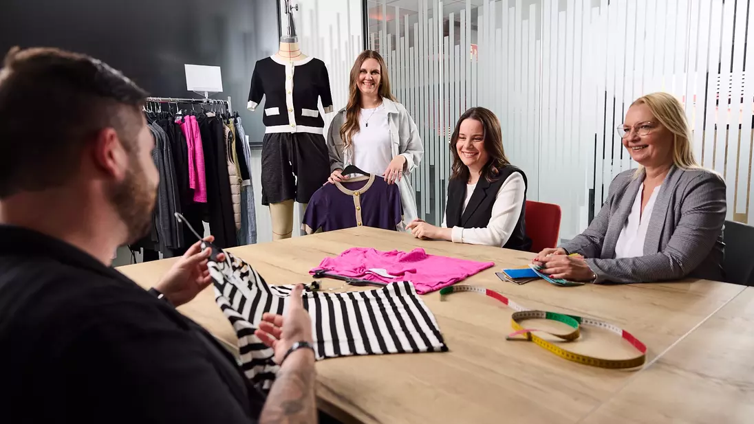 Vier Personen sitzen und stehen in einem Besprechungsraum an einem Tisch mit Kleidungsstücken, Stoffmustern und einem Maßband und besprechen eine Modekollektion.