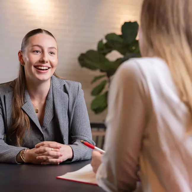 Eine Mitarbeiterin spricht mit einem lächeln mit einer anderen Mitarbeiterin an einem Tisch in einem Raum