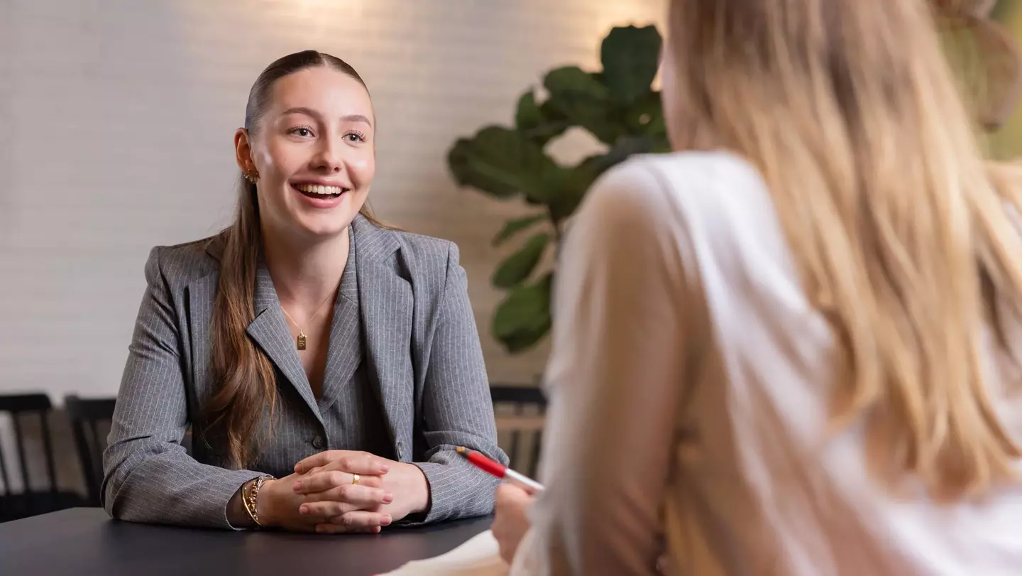 Eine Mitarbeiterin spricht mit einem lächeln mit einer anderen Mitarbeiterin an einem Tisch in einem Raum