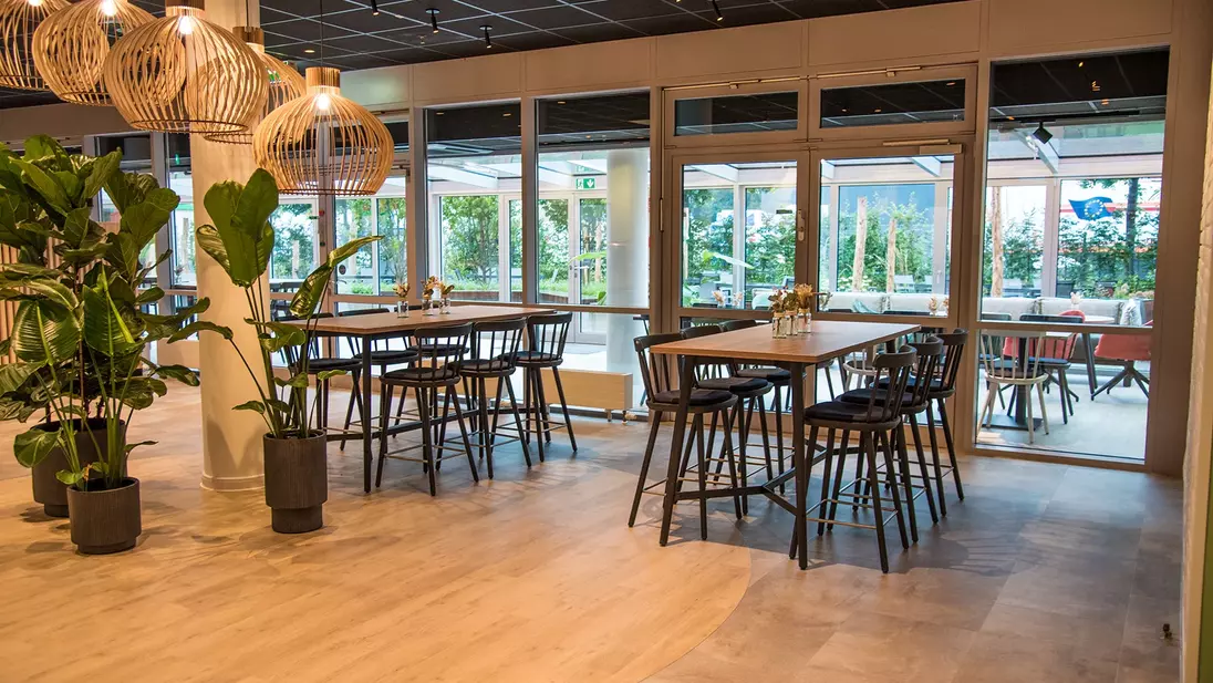 Innenansicht einer modernen Kantine mit Tischen, hohen Sitzplätzen, großen Fenstern und Pflanzen.