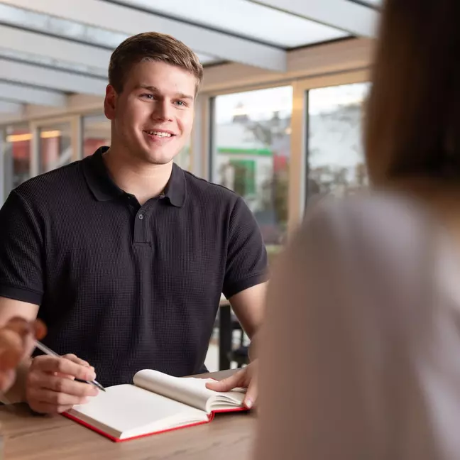 Ein Mann sitzt in einer modernen Räumlichkeit, er unterhält sich und hat ein Notizbuch geöffnet, er hält einen Stift