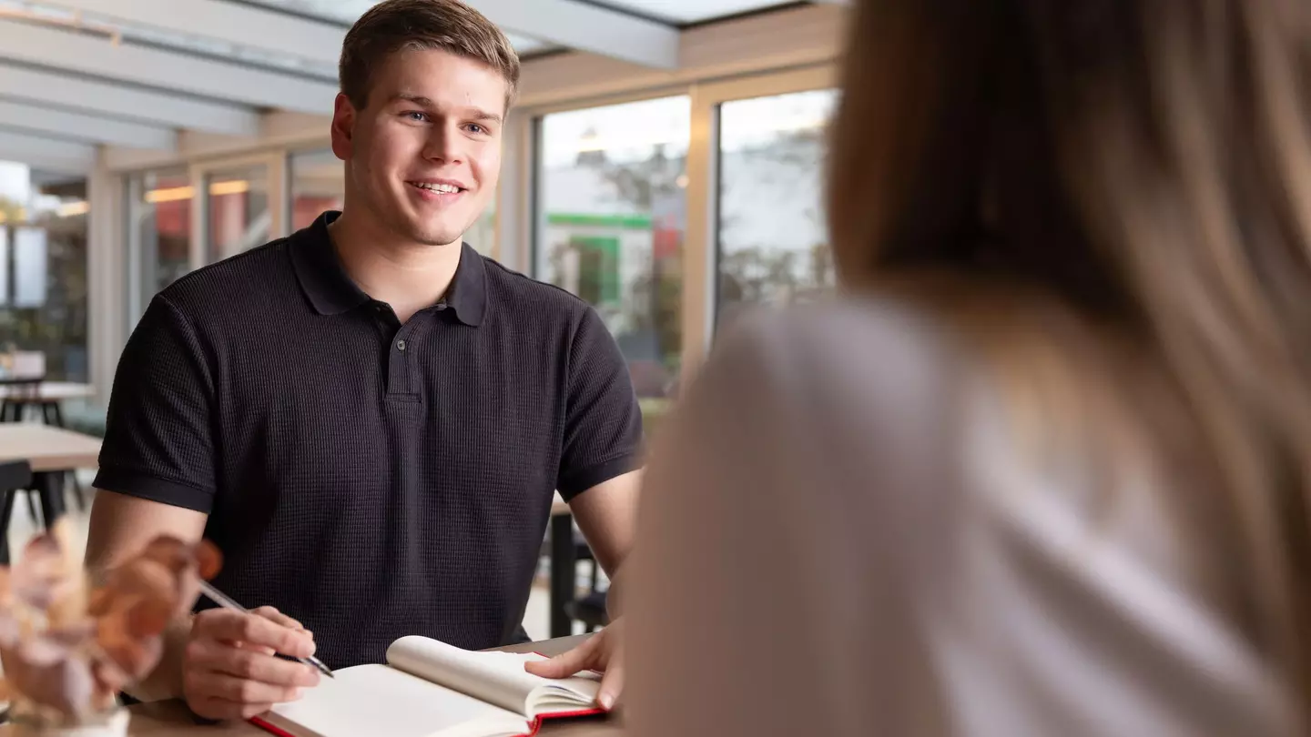 Ein Mann sitzt in einer modernen Räumlichkeit, er unterhält sich und hat ein Notizbuch geöffnet, er hält einen Stift