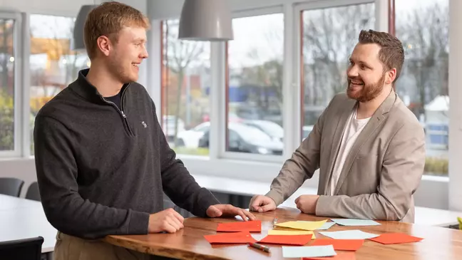 Zwei Mitarbeiter stehen lächelnd in einem modernen Büro an einem Tisch mit Post-Its