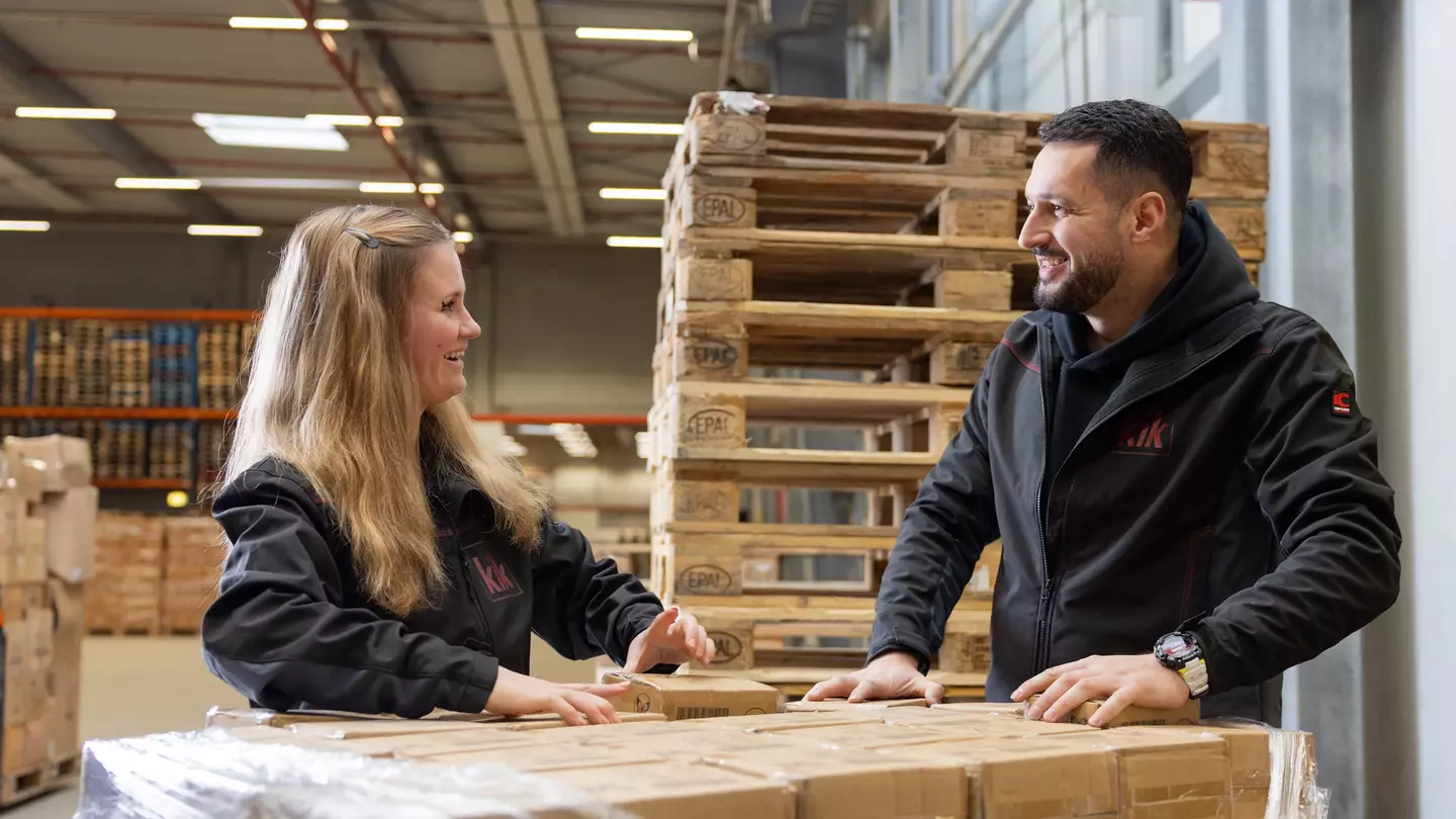 Zwei Mitarbeitende lächeln sich an, sie stehen im Lager und packen Lagerware aus