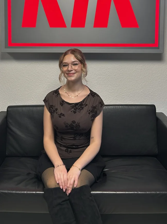 Abgebildet ist eine lächende Frau mit Brille, die auf einer schwarzen Couch sitzt.nklen Haaren.