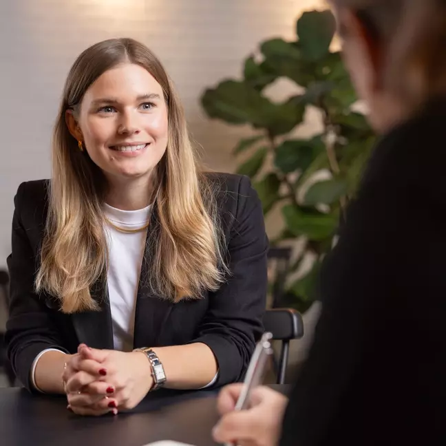 Eine Frau sitzt lächelnd an einem Tisch, sie schaut einen Mitarbeiter an