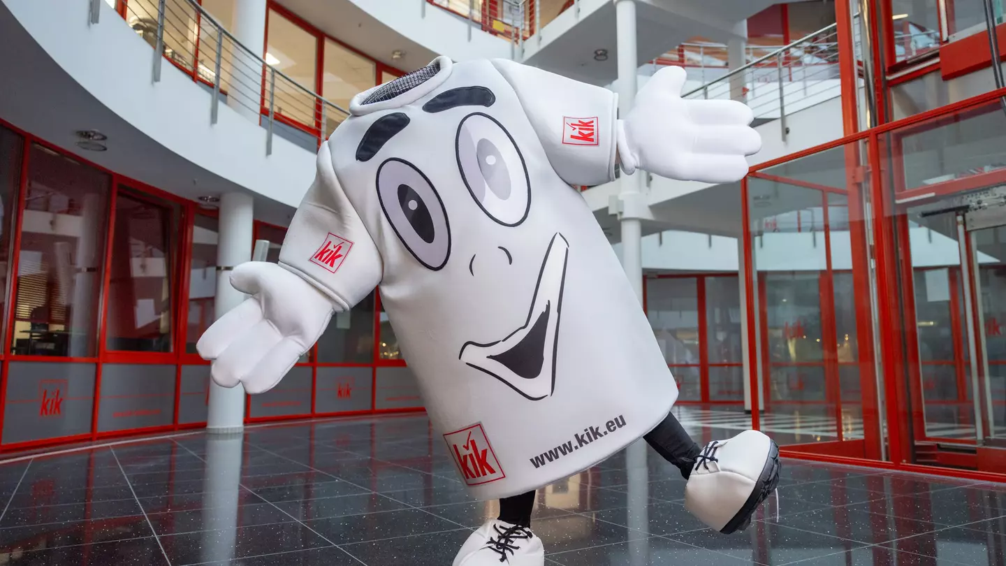 Maskottchen als großes graues T-Shirt steht in einer modernen Eingangshalle