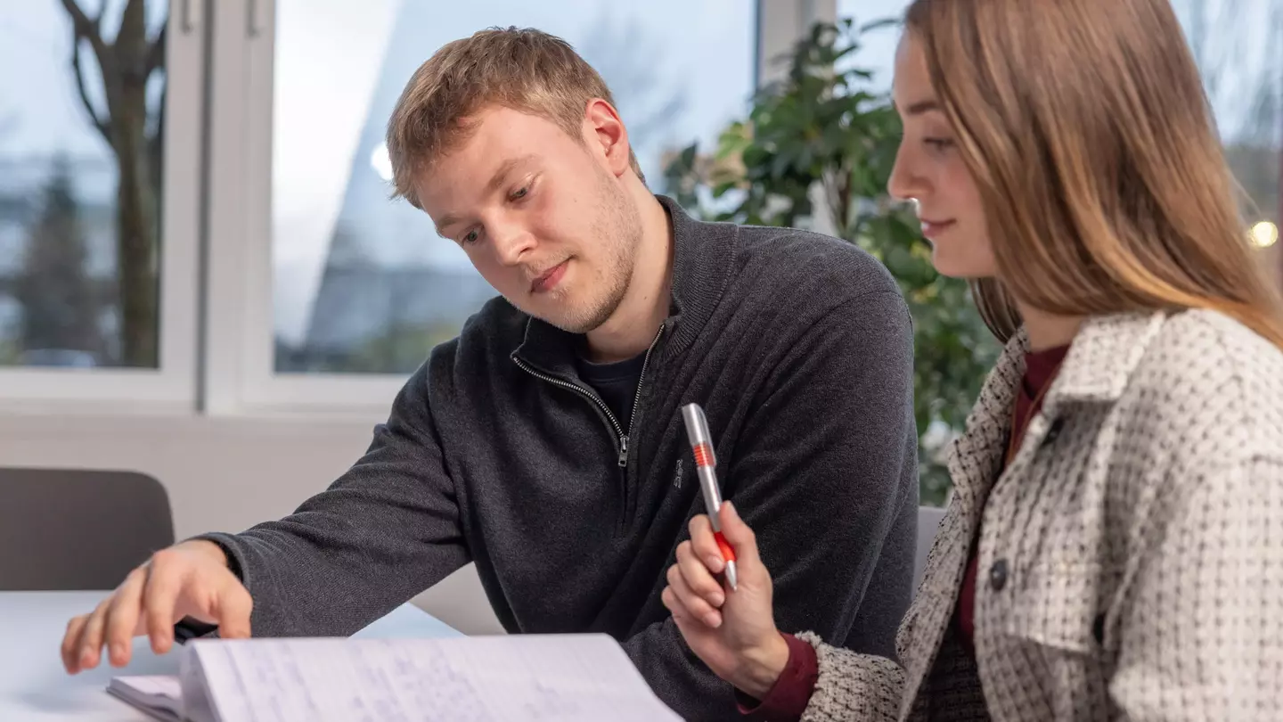 Ein Mann und eine Frau schauen in ein Notizbuch, die Frau hält einen Kugelschreiber, beide lächeln und sitzen in einem modernen Büro