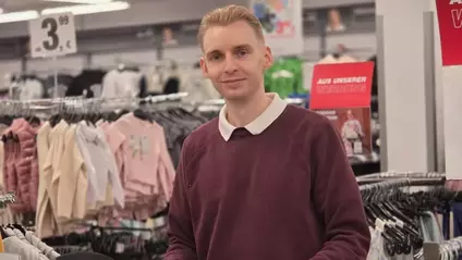 Ein Mitarbeiter im Einzelhandel steht lächelnd zwischen Kleiderständern in der Filiale.