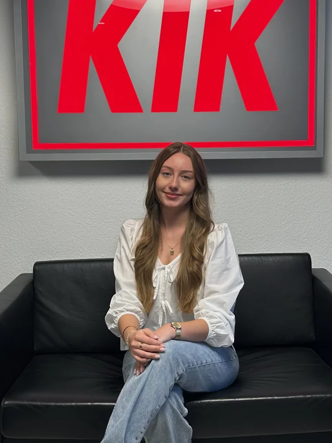 Abgebildet ist eine lächende Frau mit langen Haaren, die auf einer schwarzen Couch sitzt.