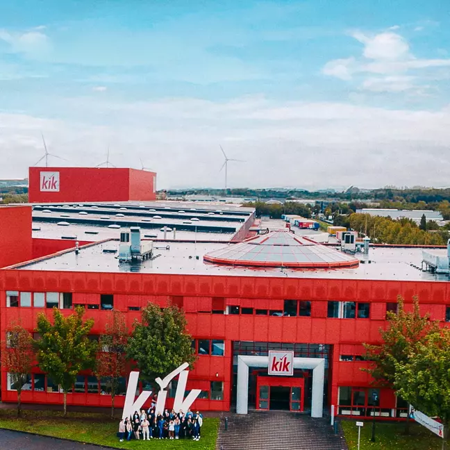 Ein großes, rotes Firmengebäude mit dem Logo „kik“ steht in einer grünen Landschaft, vor dem Eingang posiert eine Gruppe von Menschen.