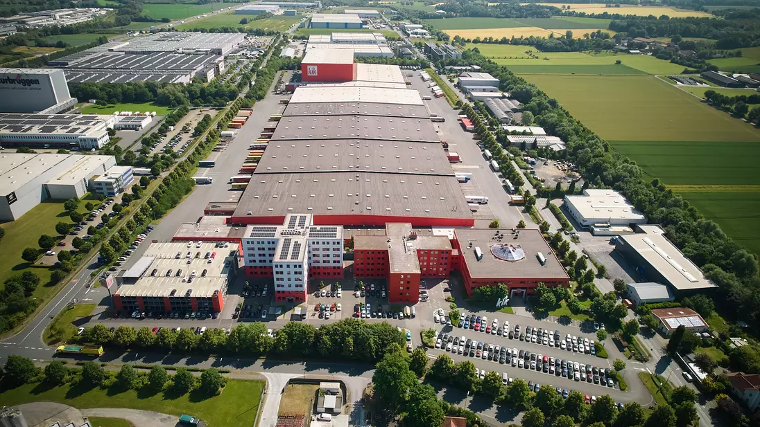 Luftaufnahme von der Zentrale mit langen Hallendächern, roten Gebäudeteilen, Parkplätzen und umliegenden Feldern.