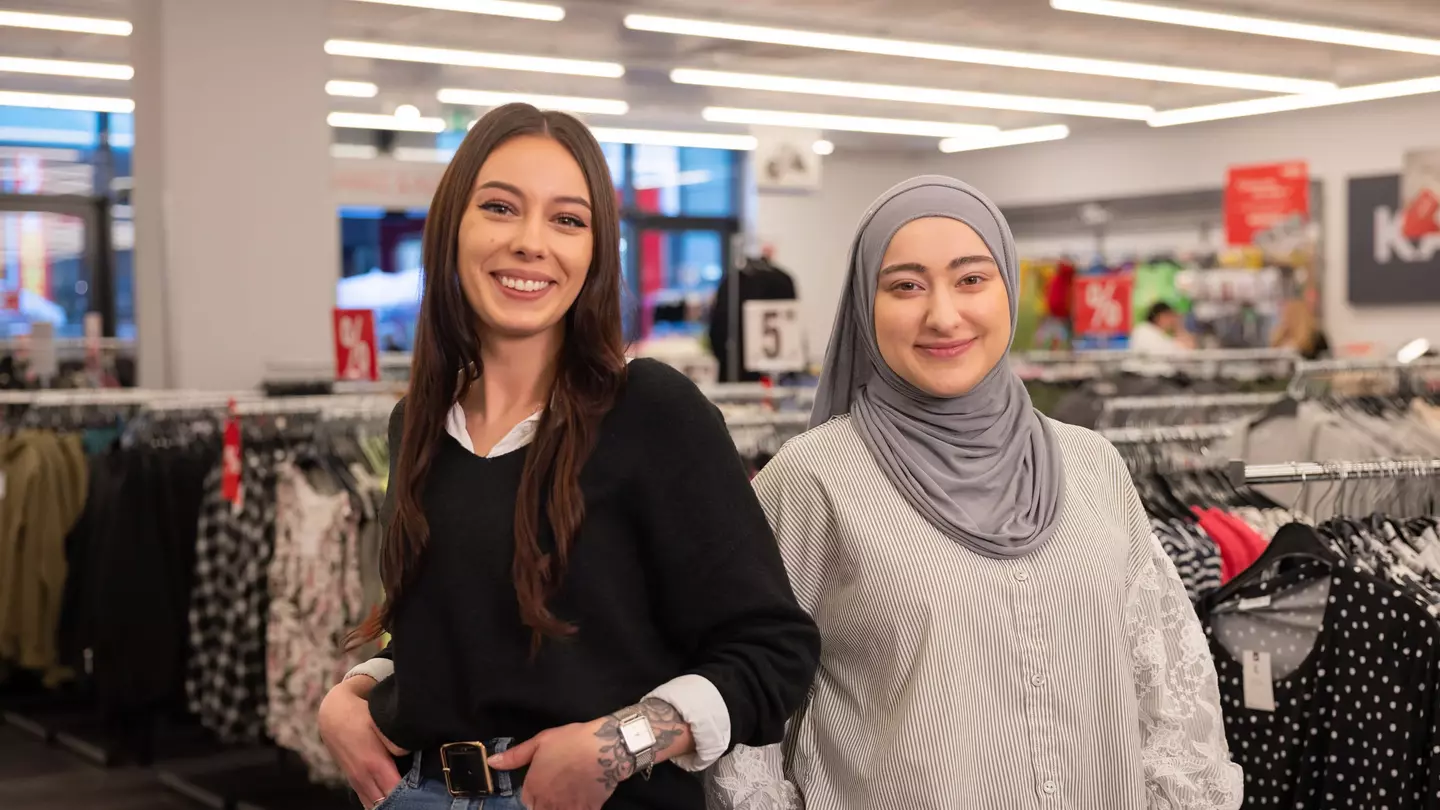 Zwei Mitarbeiterinnen im Einzelhandel stehen lächelnd zwischen Kleiderständern in der Filiale.
