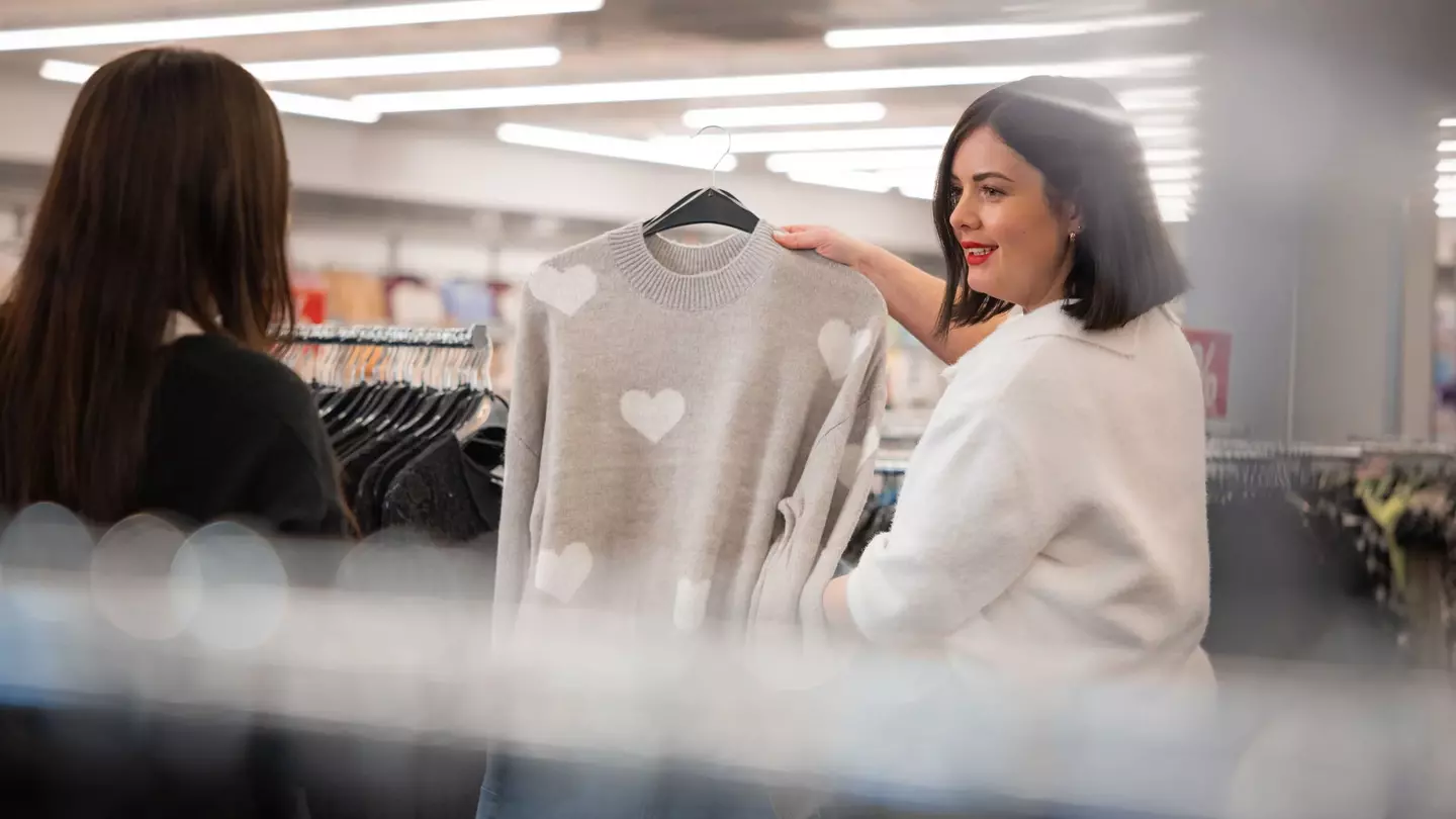 Eine Mitarbeiterin zeigt einer Kundin einen grauen Pullover mit Herzmotiv im Bekleidungsgeschäft.