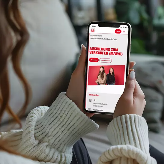 Eine Frau sitzt auf der Couch und schaut auf ihr Handydisplay, auf dem eine Stellenausschreibung von KiK angezeigt wird