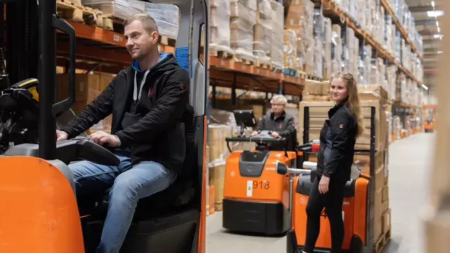 Drei Mitarbeitende arbeiten im Hochregallager, ein Mann sitzt im Gabelstapler und zwei Frauen fahren einen Hubwagen