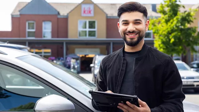 Eine Person steht vor einer KiK Filiale neben einem Auto und hält ein Tablet in der Hand.