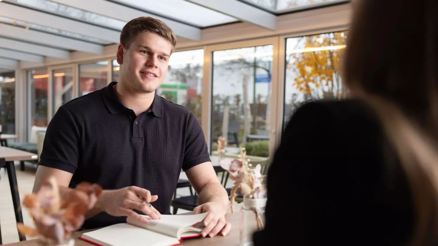 Ein Mann sitzt in einer modernen Räumlichkeit, er unterhält sich und hat ein Notizbuch geöffnet, er hält einen Stift