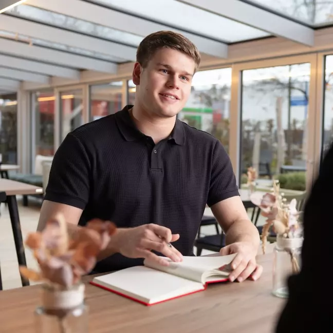 Ein Mitarbeiter sitzt mit seinem Notizbuch in der Kantine und lächelt