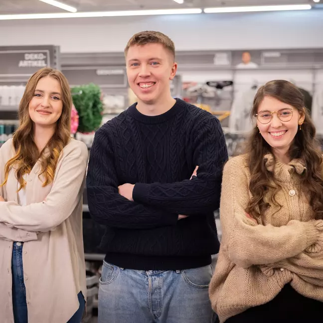 3 Mitarbeitende stehen lächelnd mit verschränkten Armen