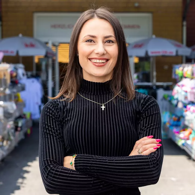 Eine Person steht im Außenbereich vor der Filiale zwischen Warenständern und blickt lächelnd in die Kamera.