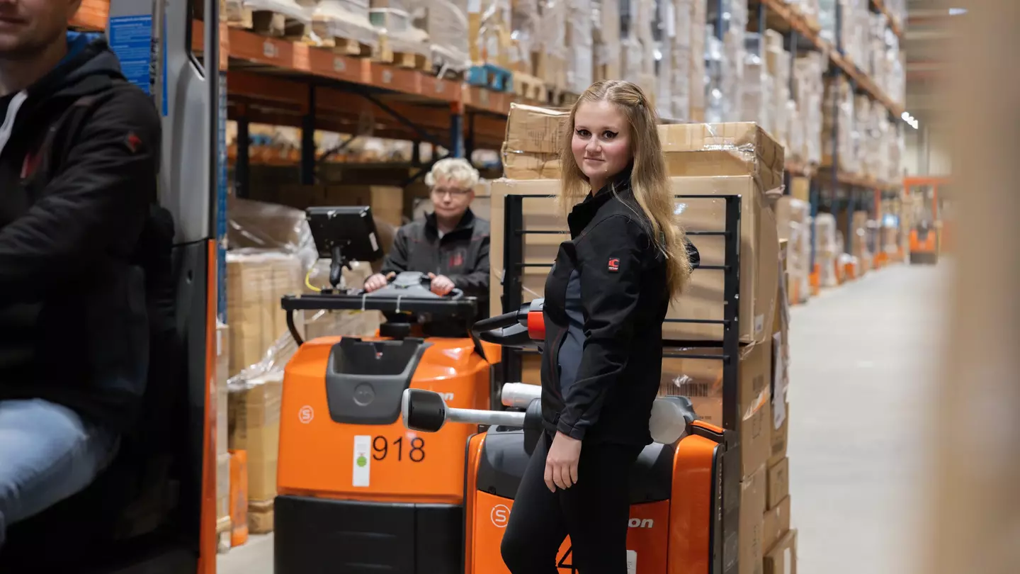 Eine Frau steht in einem Hochregallager vor einem Hubwagen welcher von einer Frau bedient wird