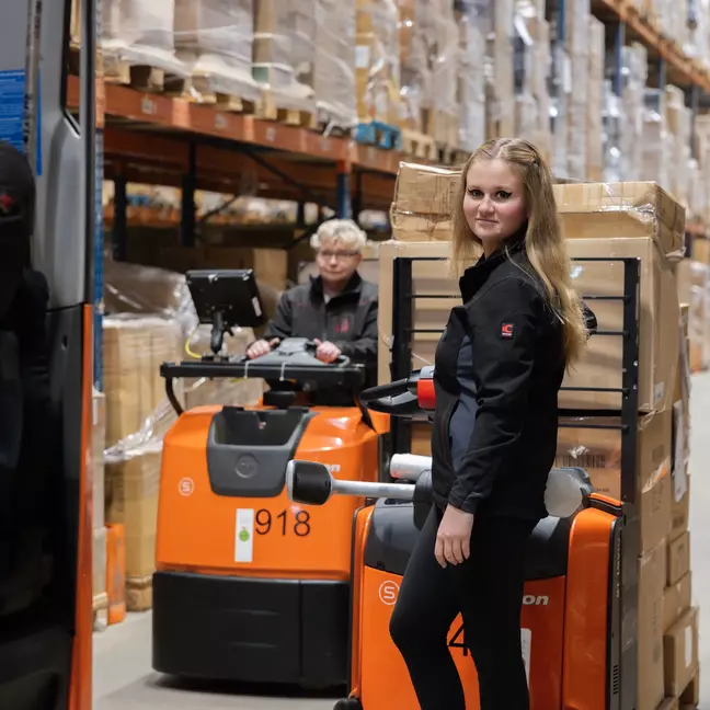 Zwei Mitarbeitende sind jeweils auf einem Gabelstabler im Lager, ein Gabelstabler ist beladen