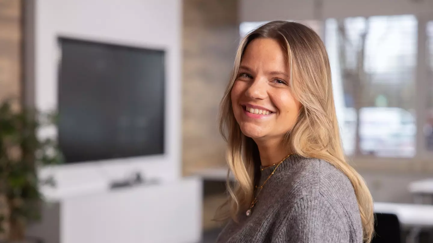 Eine Frau schaut lächelnd in die Kamera, sie steht in einer modernen Räumlichkeit