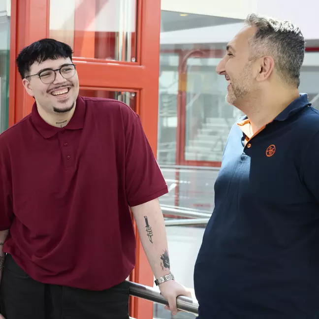 Zwei Personen stehen in einem hellen Innenbereich eines Gebäudes und unterhalten sich in entspannter Atmosphäre