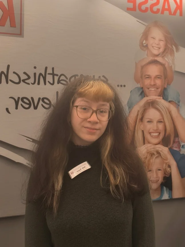 Abgebildet ist eine stehende Frau mit langen Haaren und Brille.
